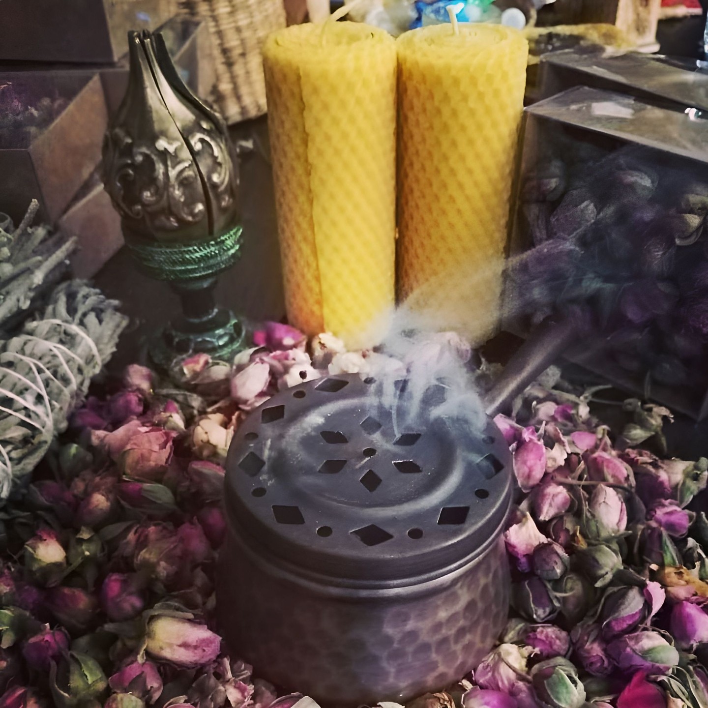 Smoke rising from a copper incense burner among dried rosebuds, sage bundles and beeswax candles